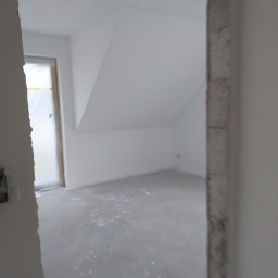 Grzegorz Bitner - Surowe wnętrze pokoju na poddaszu z widocznymi śladami po szpachlowaniu na podłodze, widoczne surowe ościeżnice drzwiowe bez zamontowanych drzwi, okno zabezpieczone folią malarską.