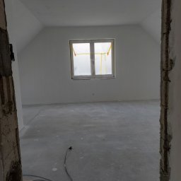 Grzegorz Bitner - Poddasze w trakcie wykańczania: świeżo pomalowane ściany na biało, okno zabezpieczone folią malarską, surowa podłoga, widoczny kabel elektryczny i fragment stojaka oświetleniowego przy ościeżnicy.