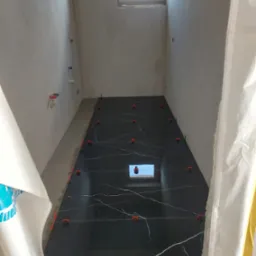 Wąskie pomieszczenie w trakcie układania podłogi z czarnych płytek imitujących marmur, z użyciem pomarańczowych klipsów poziomujących; widoczne surowe ściany i okno w górnej części ściany.