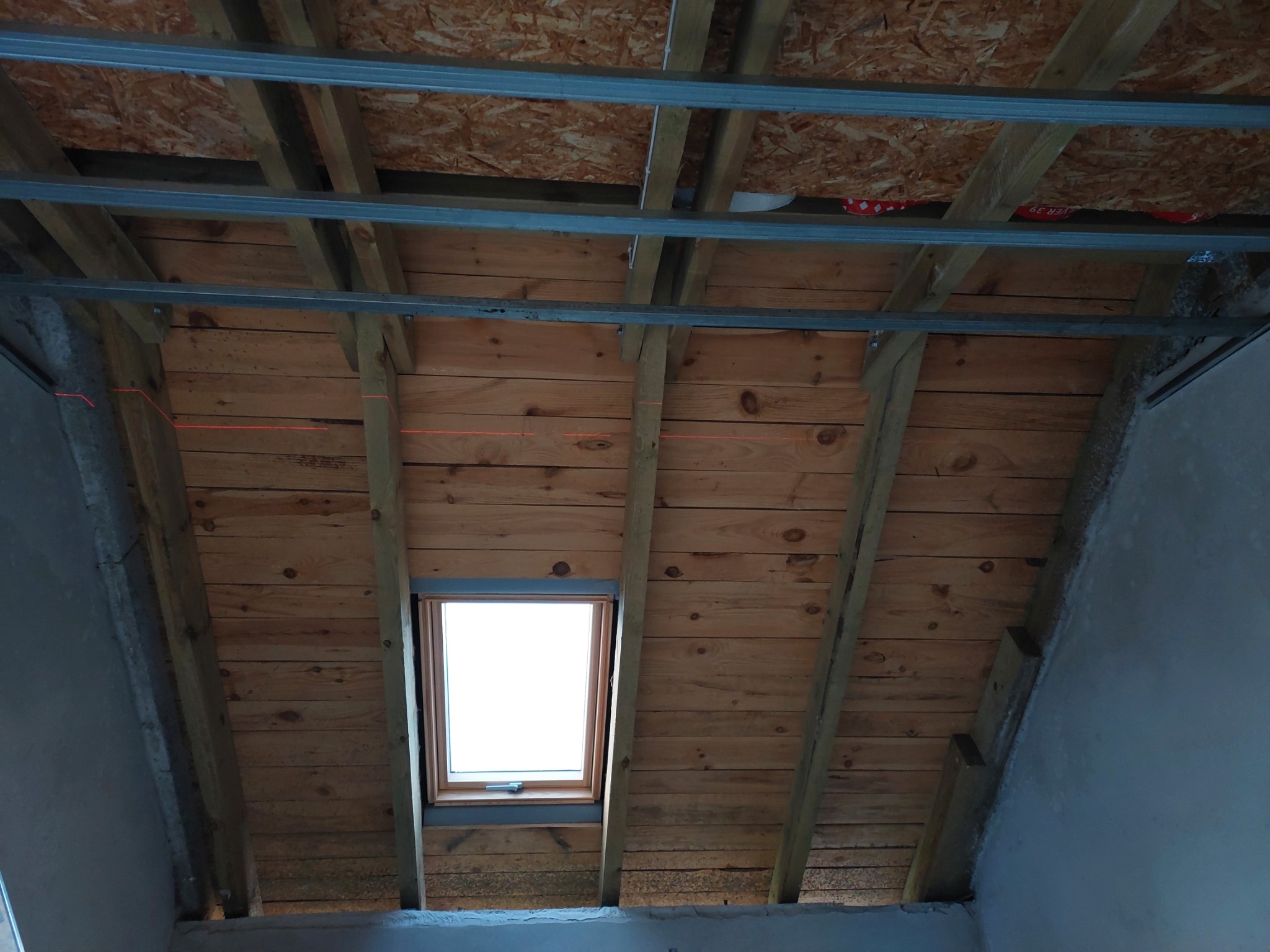 Poddasze w trakcie adaptacji: drewniana konstrukcja dachu z oknem połaciowym, metalowe profile sufitowe zamontowane, ściany przygotowane do wykończenia, widoczne ślady po laserowym wyznaczaniu linii.