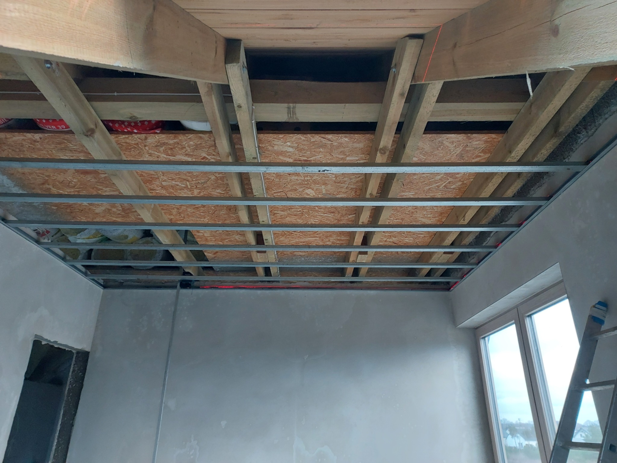 Konstrukcja sufitu podwieszanego z metalowych profili, widoczne drewniane elementy stropu i płyta OSB, surowe ściany w trakcie wykończenia, drabina oparta o ścianę przy oknie.