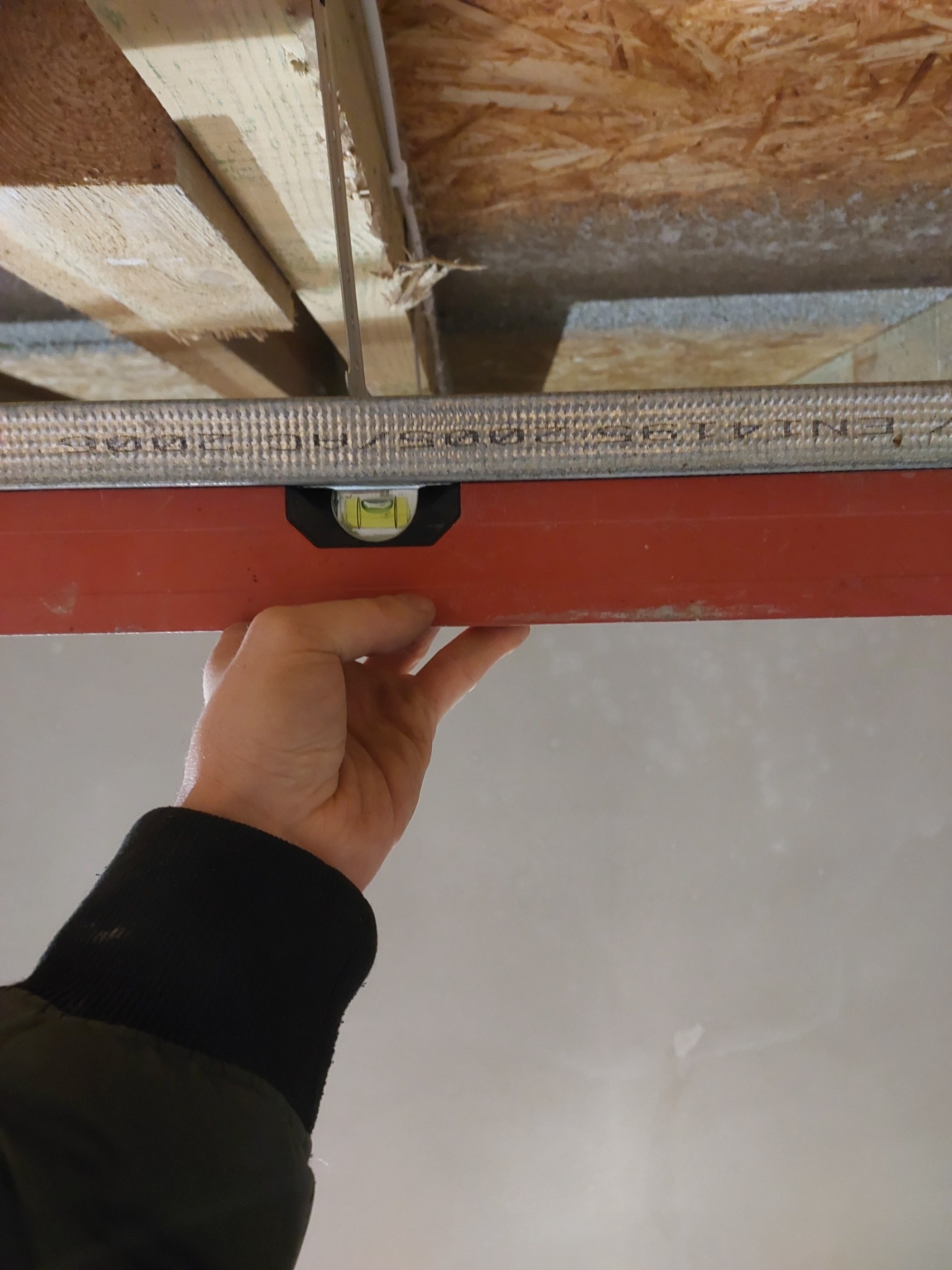 Ręka przytrzymuje poziomicę na metalowym profilu pod drewnianym stropem, widoczny fragment sufitu w trakcie prac wykończeniowych.