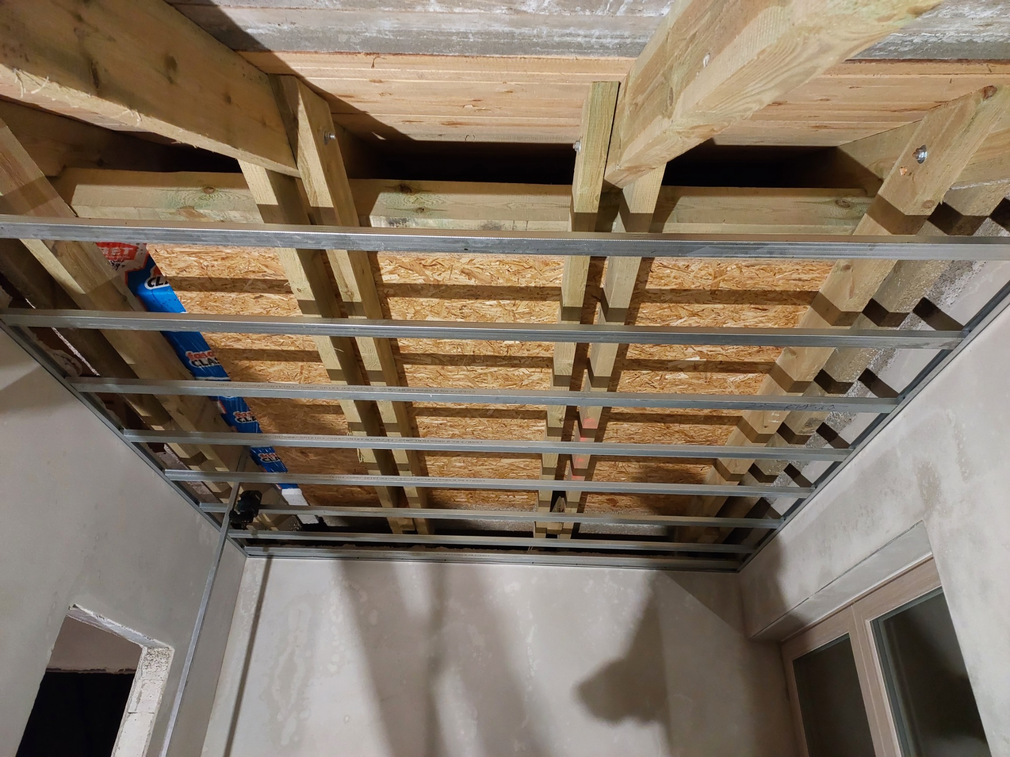 Sufit podwieszany w trakcie montażu: drewniana konstrukcja nośna, metalowy stelaż z profili, płyta OSB, widoczne ściany w trakcie wykańczania gładzią.