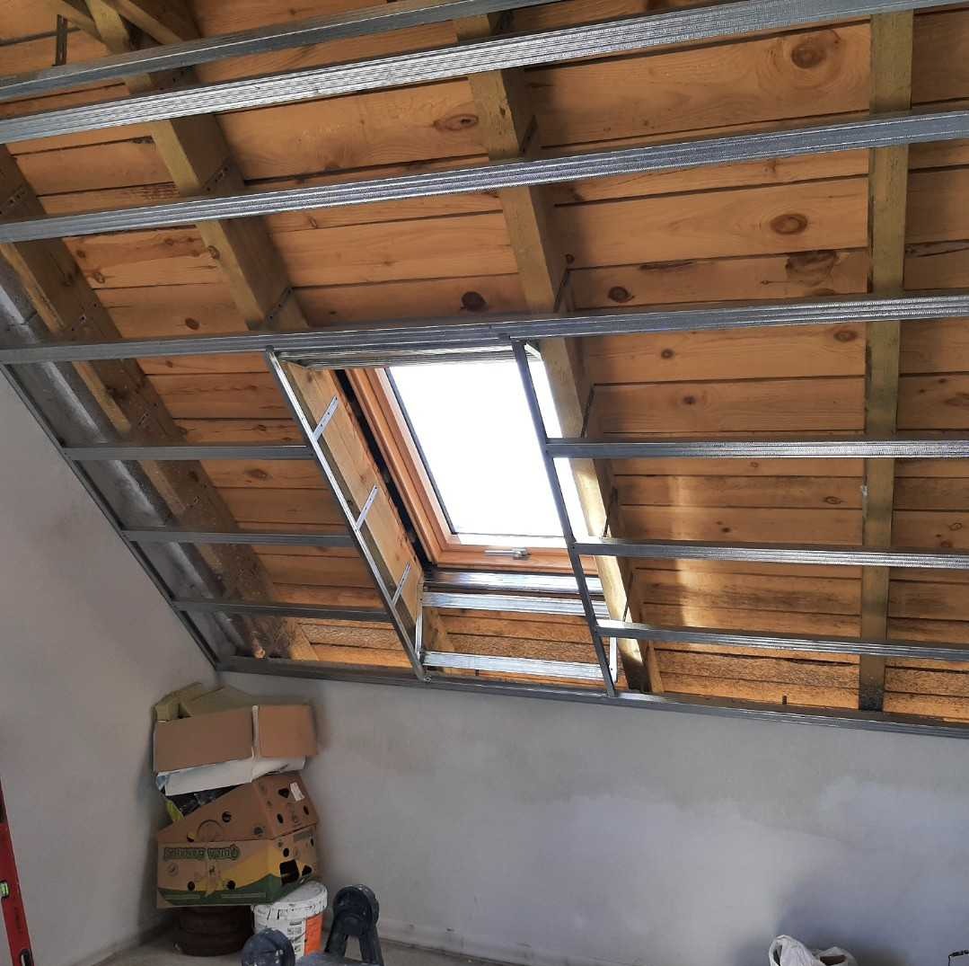 Konstrukcja metalowa pod płytę gipsowo-kartonową na drewnianym poddaszu z oknem dachowym, widoczne kartony i narzędzia na podłodze.