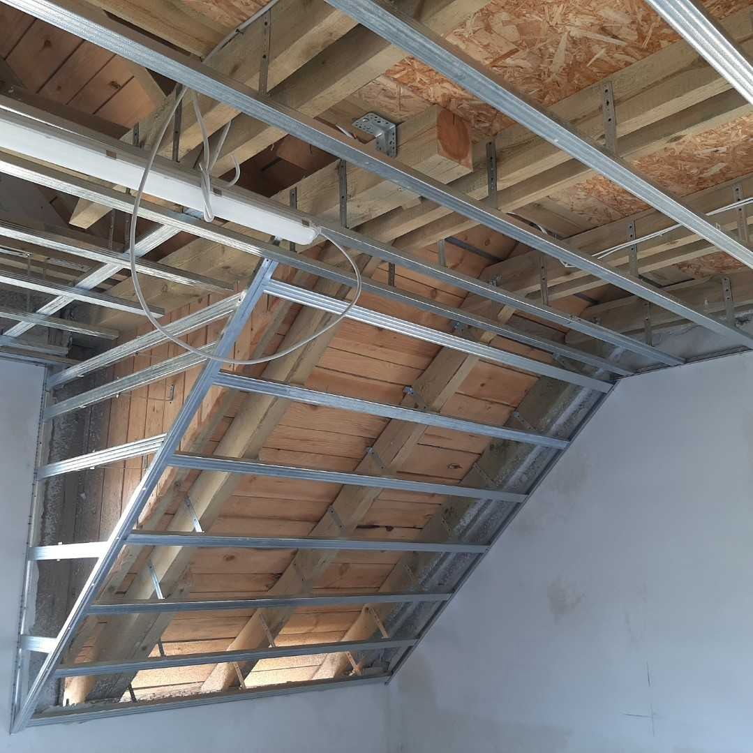 Konstrukcja metalowa podwieszanego sufitu z widocznymi drewnianymi belkami stropu i fragmentem ściany w trakcie prac wykończeniowych.