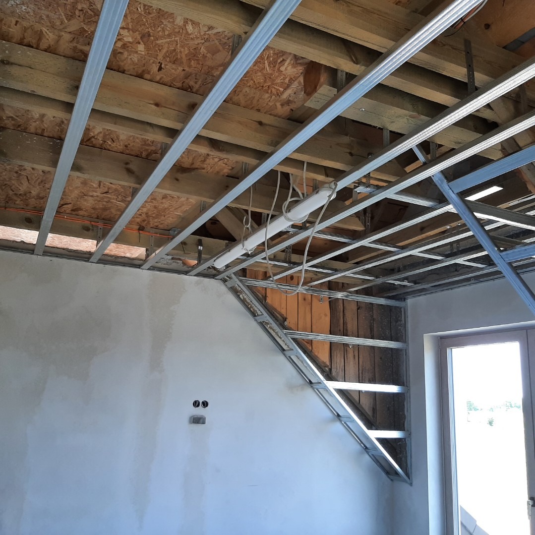 Wnętrze w trakcie montażu stelaża z profili metalowych pod płytę gipsowo-kartonową na skośnej ścianie poddasza z widoczną drewnianą konstrukcją dachu i instalacją elektryczną.