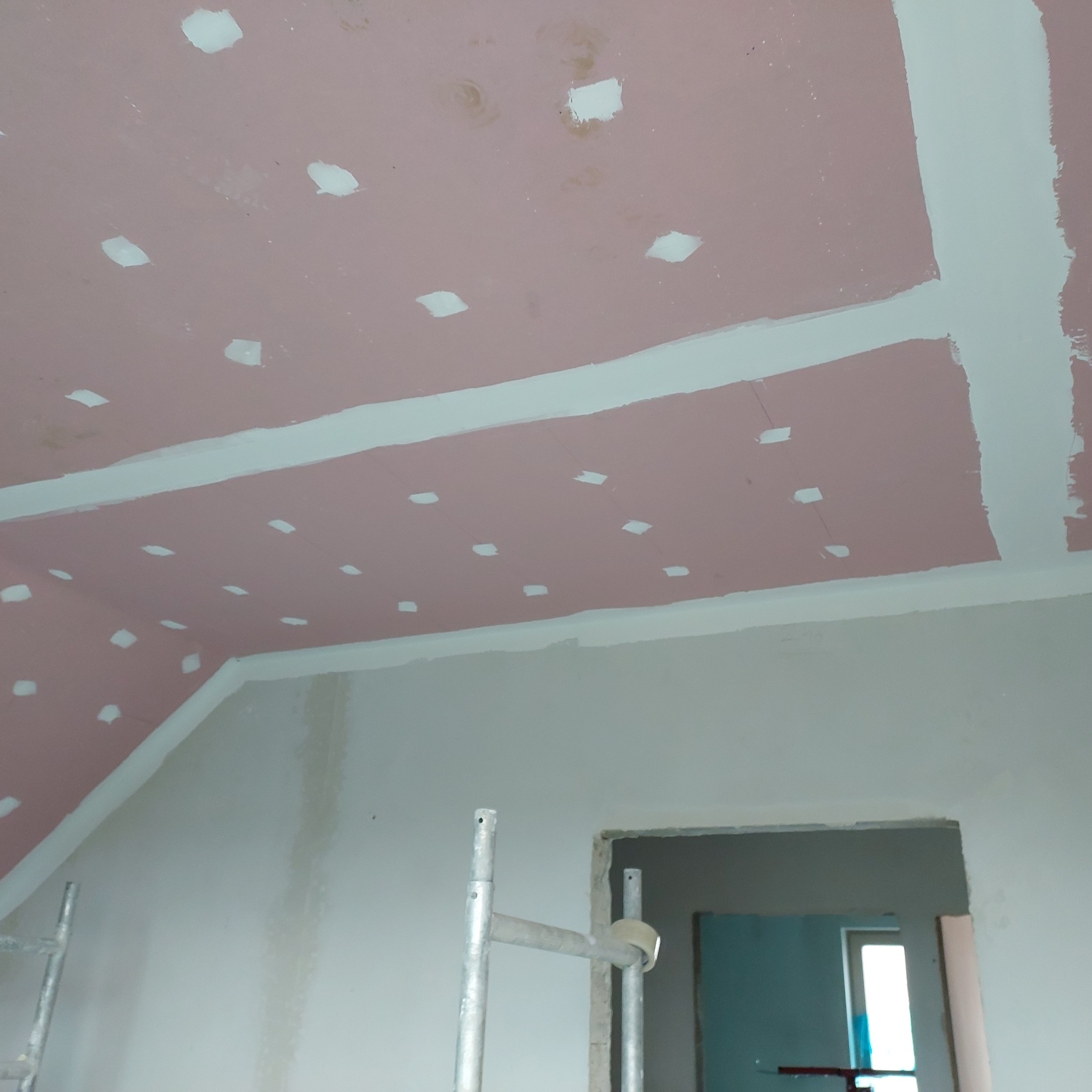 Różowa płyta kartonowo-gipsowa na suficie z widocznymi białymi szpachlowanymi punktami mocowań, białe łączenia płyt i fragment rusztowania w pomieszczeniu w trakcie remontu.
