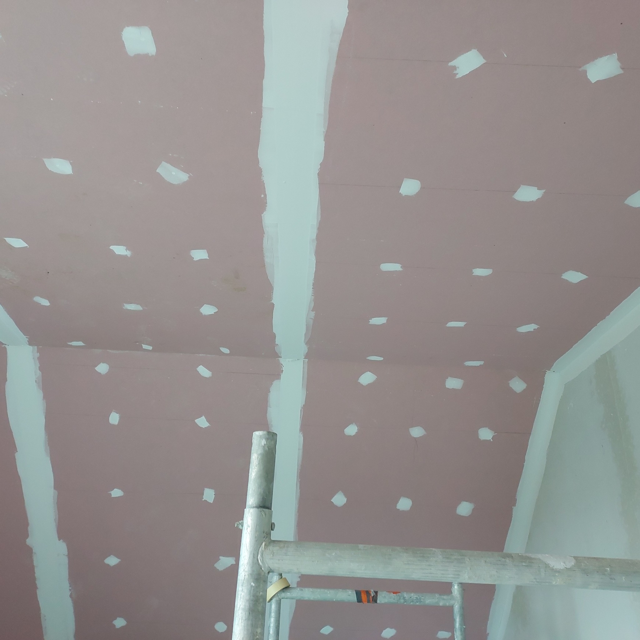 Sufit z płyt kartonowo-gipsowych, wstępnie szpachlowany, z widoczną siatką spoin i miejscami po wkrętach, częściowo zakryty przez metalowe rusztowanie.