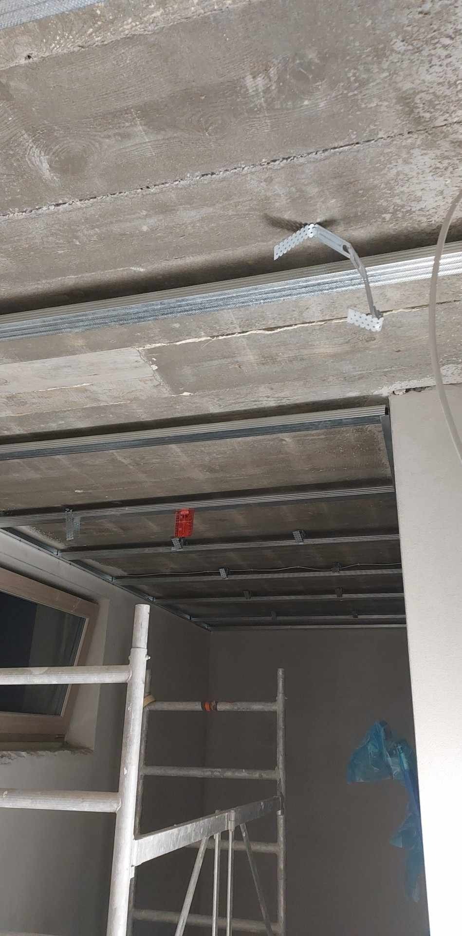 Montaż metalowego rusztu pod płytę karton gips na surowym betonowym suficie, widoczne profile i uchwyty montażowe, rusztowanie malarskie w pomieszczeniu.
