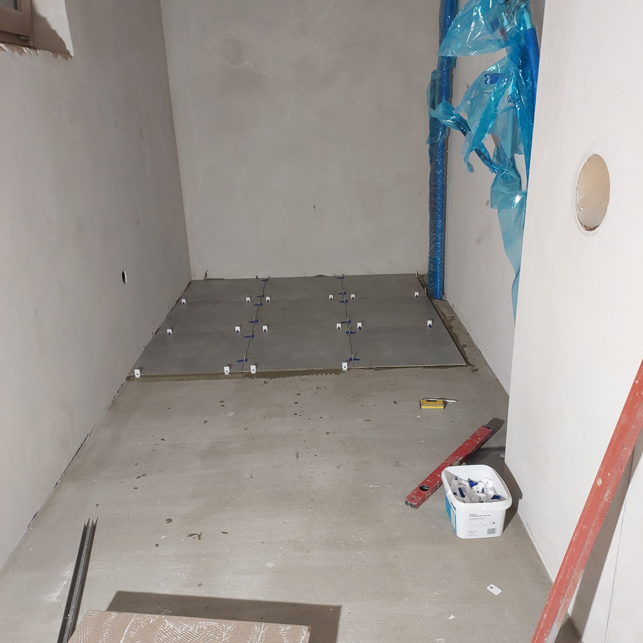 Układanie szarych płytek podłogowych z użyciem systemu poziomowania, widoczne narzędzia i poziomice, ściany przygotowane do dalszych prac wykończeniowych, rury zabezpieczone folią.