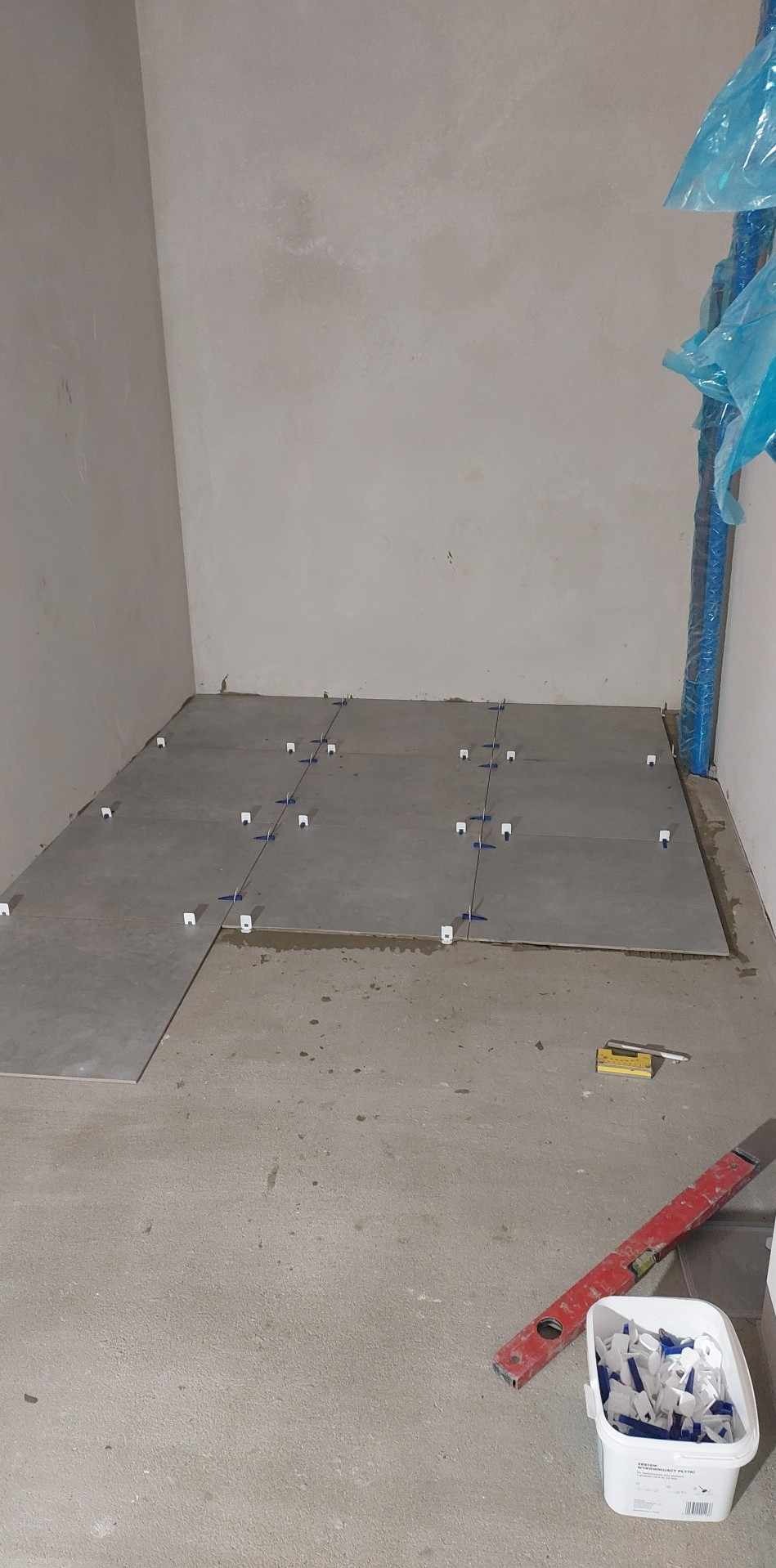 Układanie szarych płytek podłogowych w surowym wnętrzu, z użyciem białych klipsów poziomujących i czerwonej poziomicy, rury zabezpieczone folią.