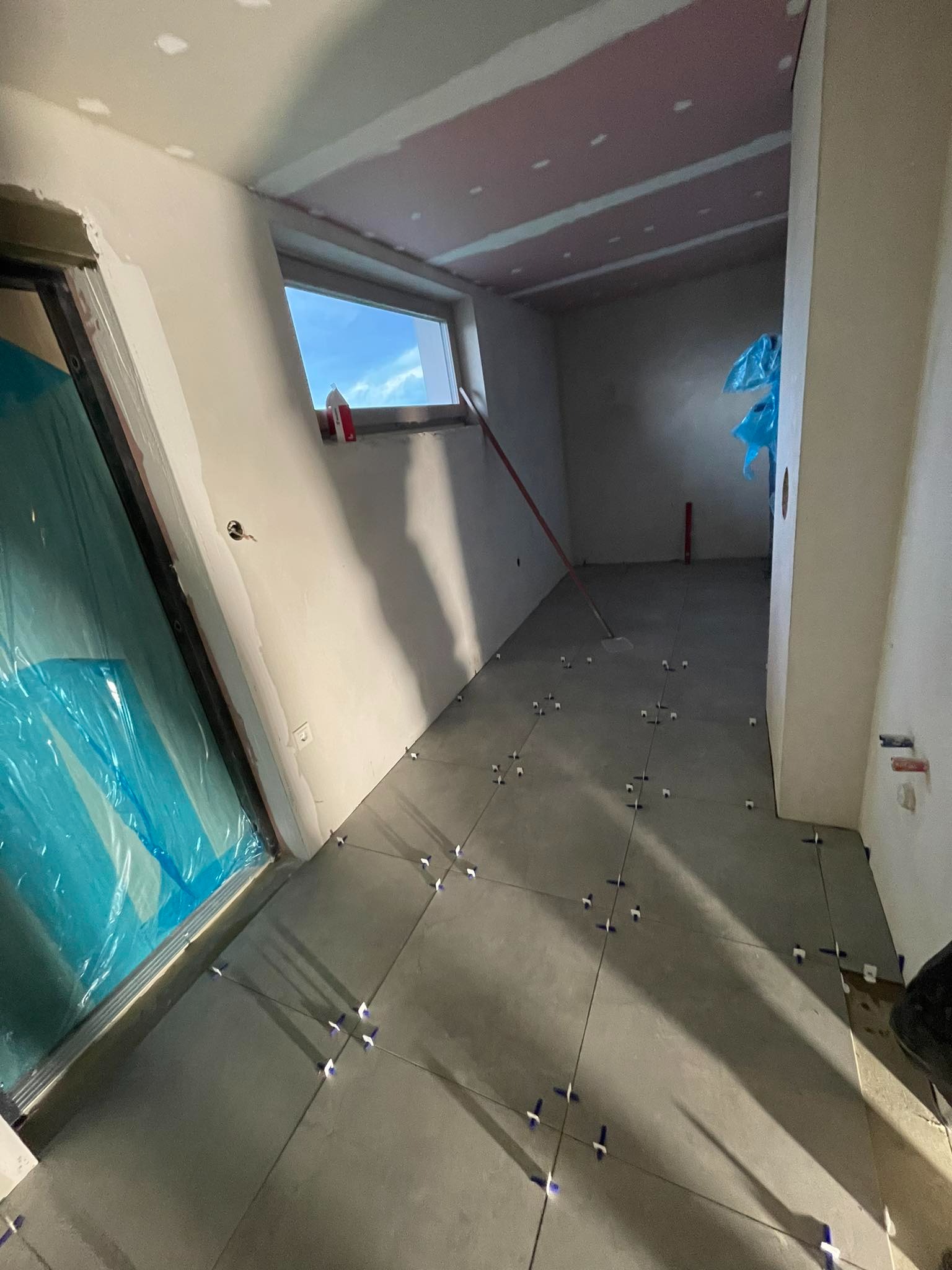 Wnętrze w trakcie remontu: szare płytki podłogowe z krzyżykami dystansowymi, ściany przygotowane do malowania, okno z widokiem na niebo, zabezpieczone folią drzwi.