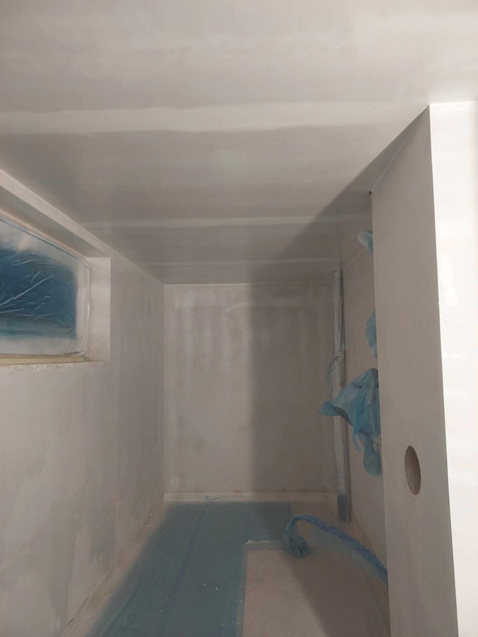 Przygotowane do malowania pomieszczenie z zabezpieczonym oknem i podłogą folią malarską, ściany po szpachlowaniu, widoczny fragment rury również zabezpieczony folią.