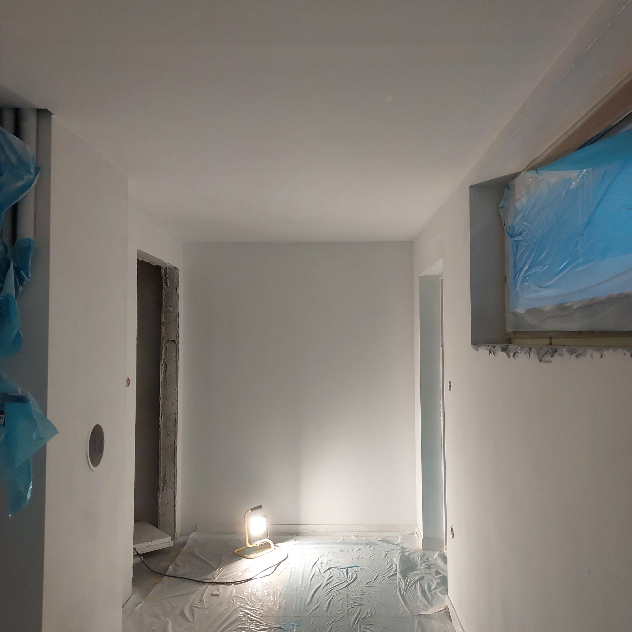 Wnętrze po świeżym malowaniu na biało, z zabezpieczonymi folią oknami i podłogą, oświetlone lampą roboczą.
