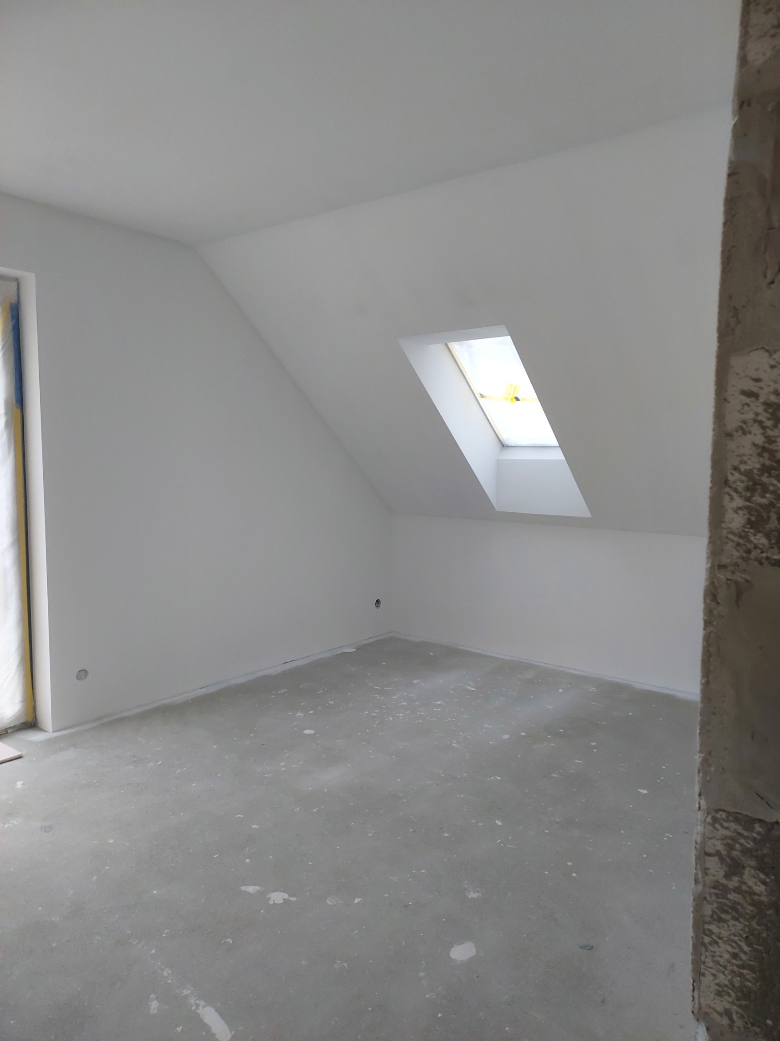 Poddasze po szpachlowaniu i malowaniu na biało, widoczne ślady po pracach na podłodze, okno dachowe w skosie dachu, surowy filar po prawej stronie kadru.