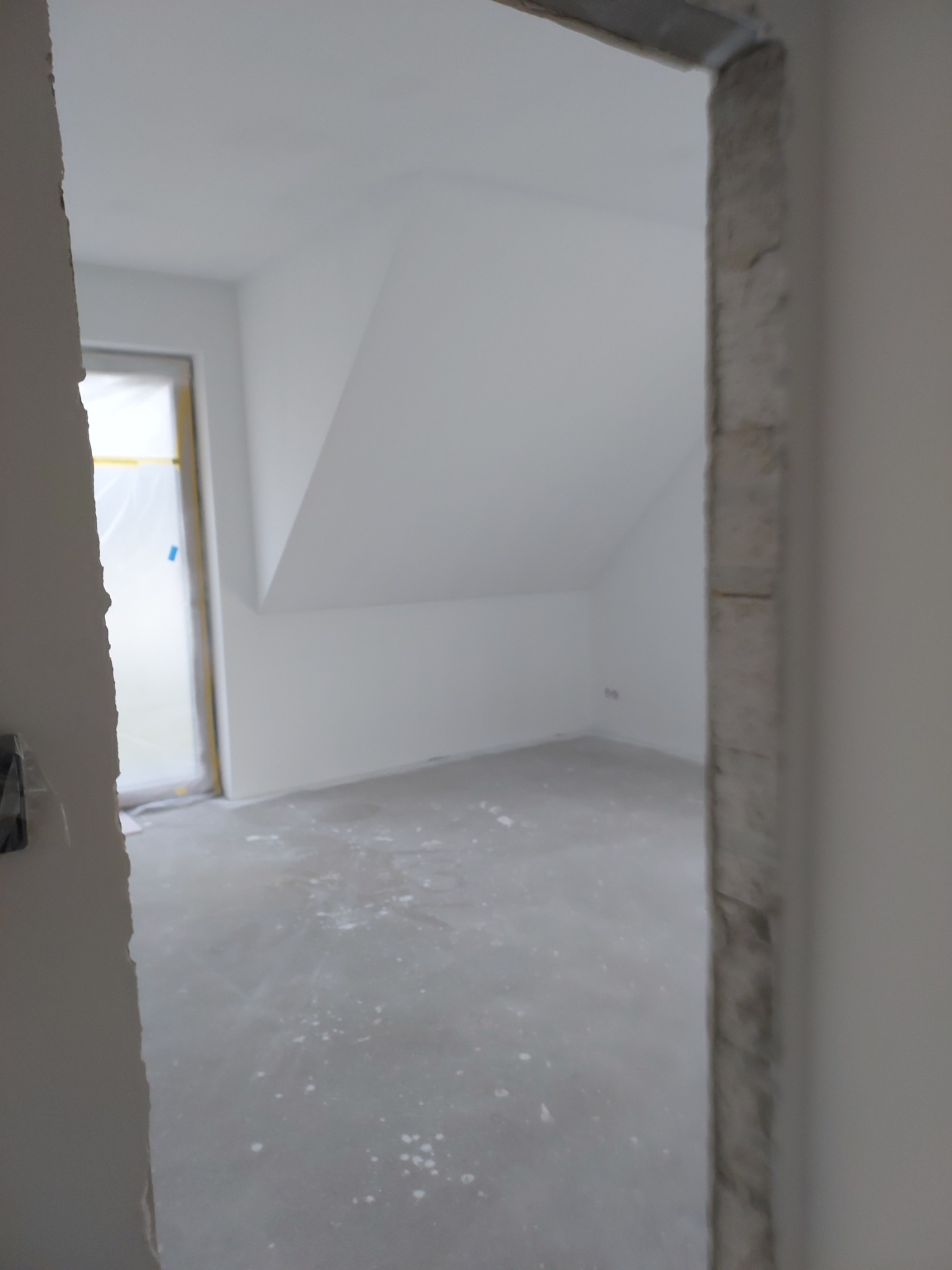 Surowe wnętrze pokoju na poddaszu z widocznymi śladami po szpachlowaniu na podłodze, widoczne surowe ościeżnice drzwiowe bez zamontowanych drzwi, okno zabezpieczone folią malarską.