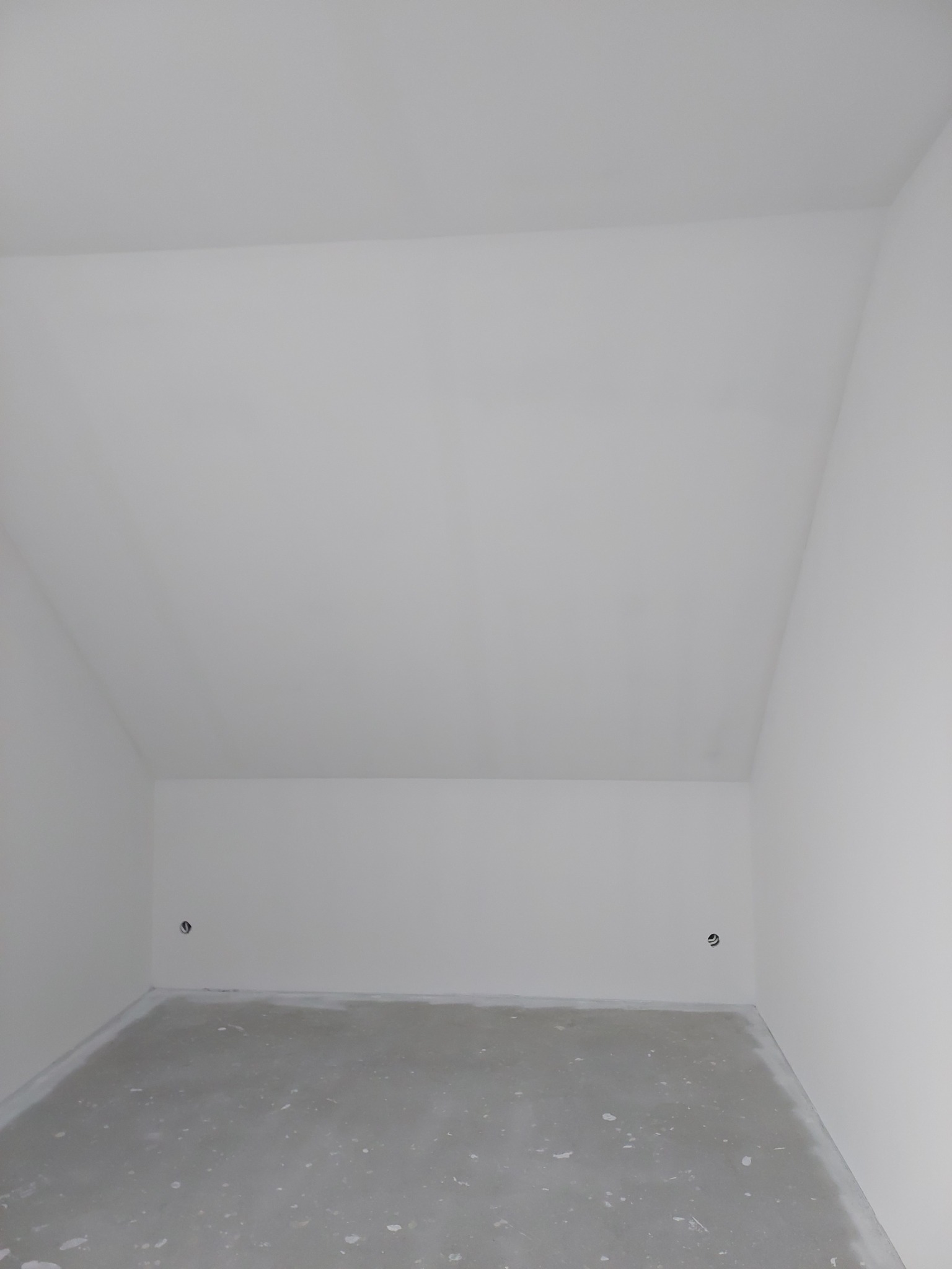 Poddasze po szpachlowaniu i gruntowaniu ścian, widoczne gniazdka elektryczne, surowy beton na podłodze, przygotowane do dalszych prac wykończeniowych.