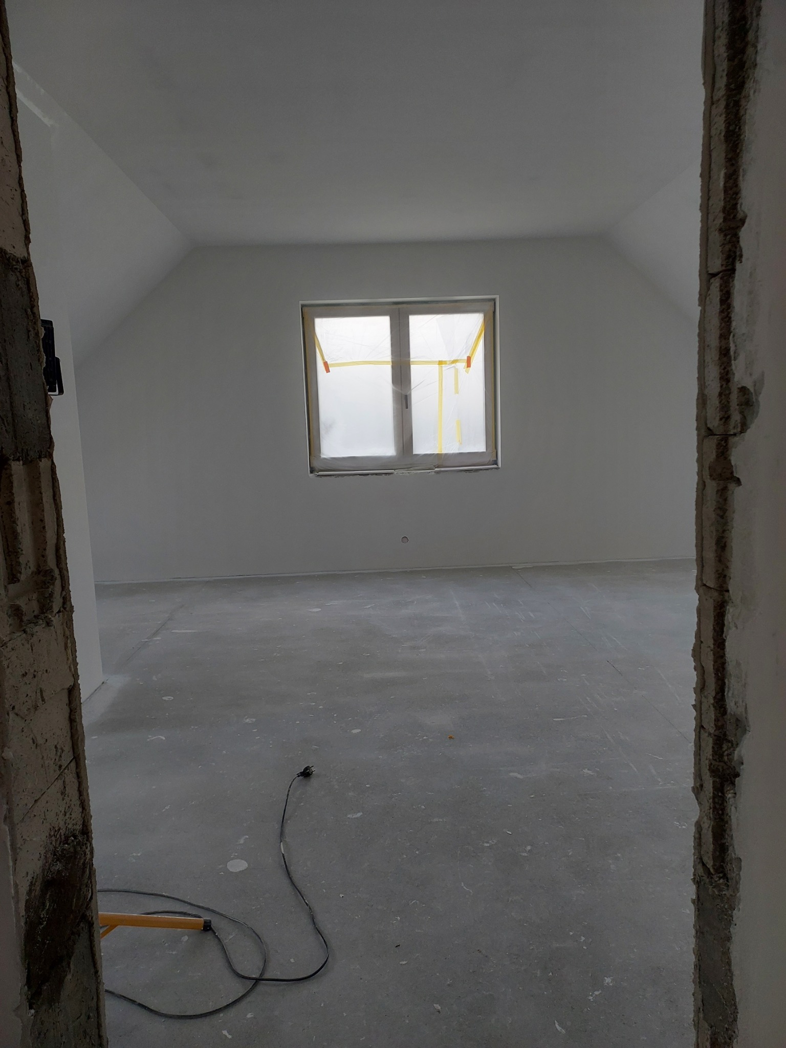 Poddasze w trakcie wykańczania: świeżo pomalowane ściany na biało, okno zabezpieczone folią malarską, surowa podłoga, widoczny kabel elektryczny i fragment stojaka oświetleniowego przy ościeżnicy.