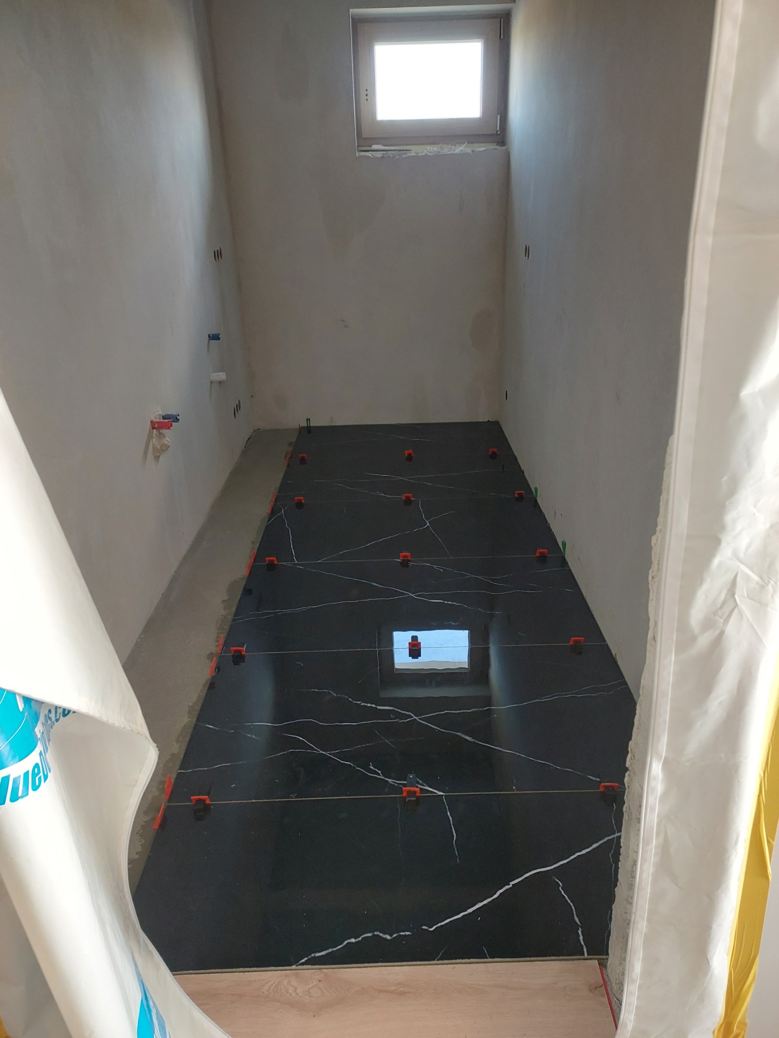 Wąskie pomieszczenie w trakcie układania podłogi z czarnych płytek imitujących marmur, z użyciem pomarańczowych klipsów poziomujących; widoczne surowe ściany i okno w górnej części ściany.