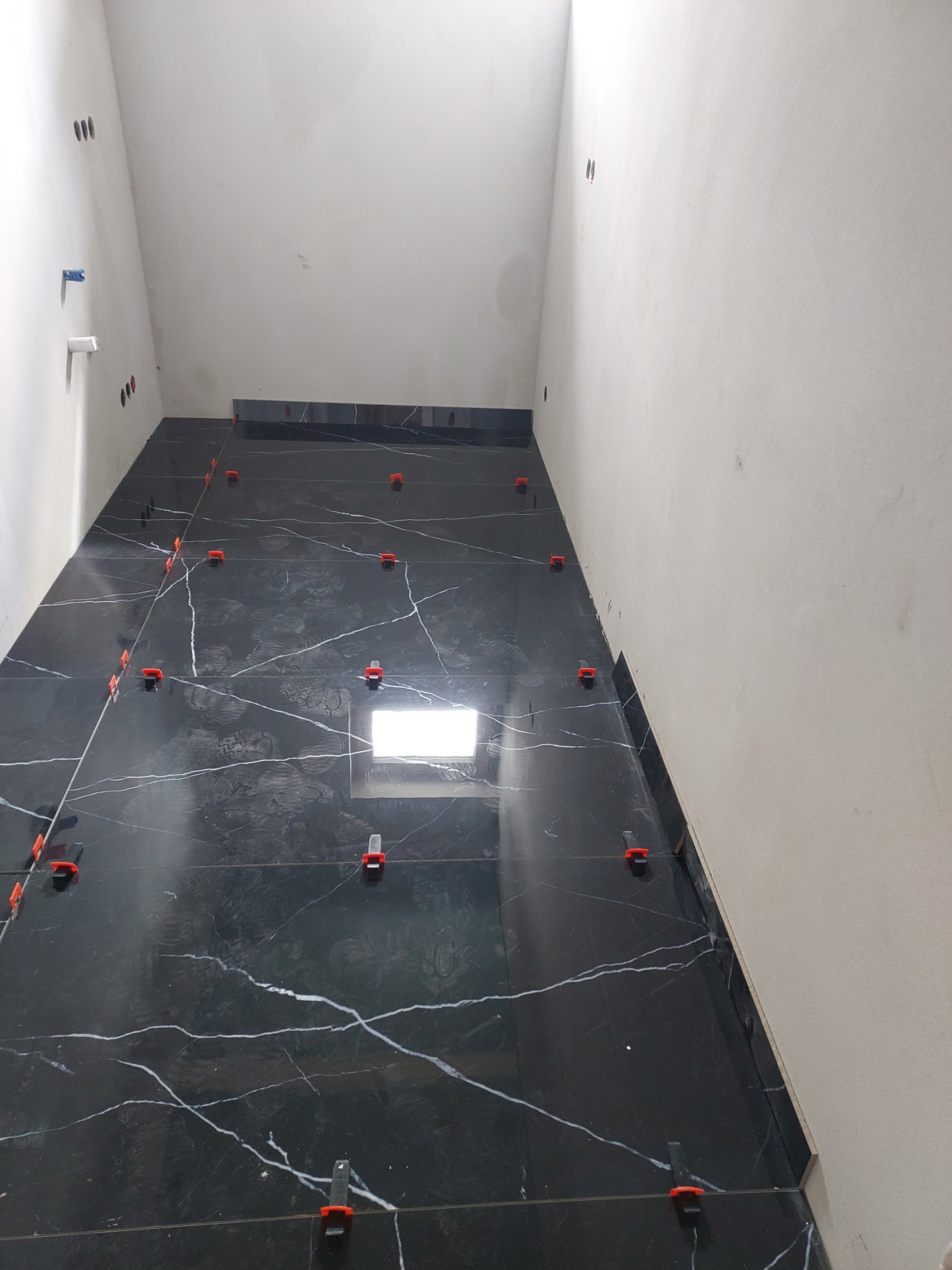 Świeżo ułożone, czarne płytki imitujące marmur z białymi żyłami, z pomarańczowymi klipsami poziomującymi na ścianie w trakcie remontu łazienki.