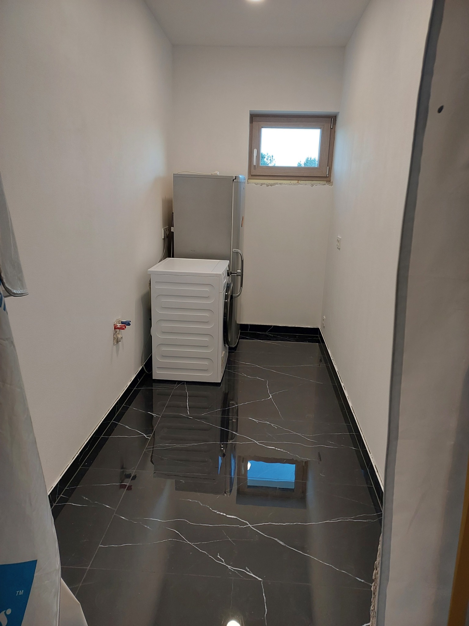 Wnętrze małej łazienki z czarną marmurową podłogą, białą pralką i lodówką, okno z drewnianą ramą w tle, białe ściany.