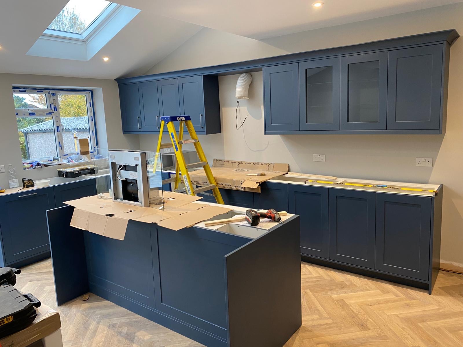 Montaż niebieskiej zabudowy kuchennej z wyspą na parkiecie w jodełkę. Widoczna drabina, narzędzia i fragment okapu. Oświetlenie sufitowe i okno dachowe.