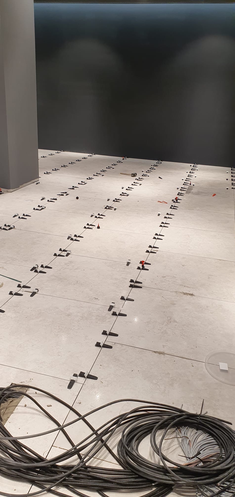 Układanie jasnych płytek podłogowych z użyciem klipsów poziomujących. Widoczne przewody w dolnej części kadru i szara ściana w tle.