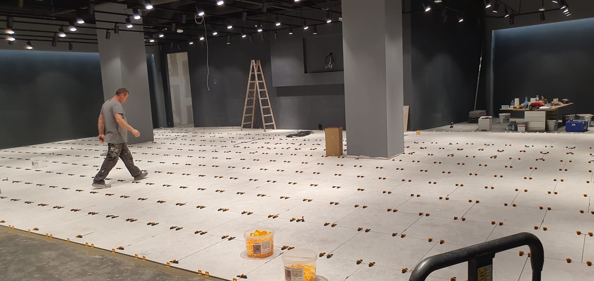 Układanie jasnoszarej podłogi z płytek w lokalu komercyjnym. Widoczny glazurnik podczas pracy, poziomica, drabina oraz pojemniki z elementami do poziomowania płytek.