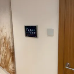 Panel sterowania inteligentnym domem zamontowany na ścianie, obok obrazu i drzwi z drewnianą okleiną.