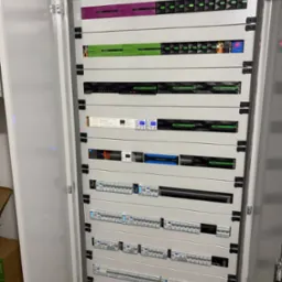 Otwarta szafa sterownicza z modułami Loxone, bezpiecznikami i innymi urządzeniami, widoczne diody sygnalizacyjne i wyświetlacze cyfrowe.