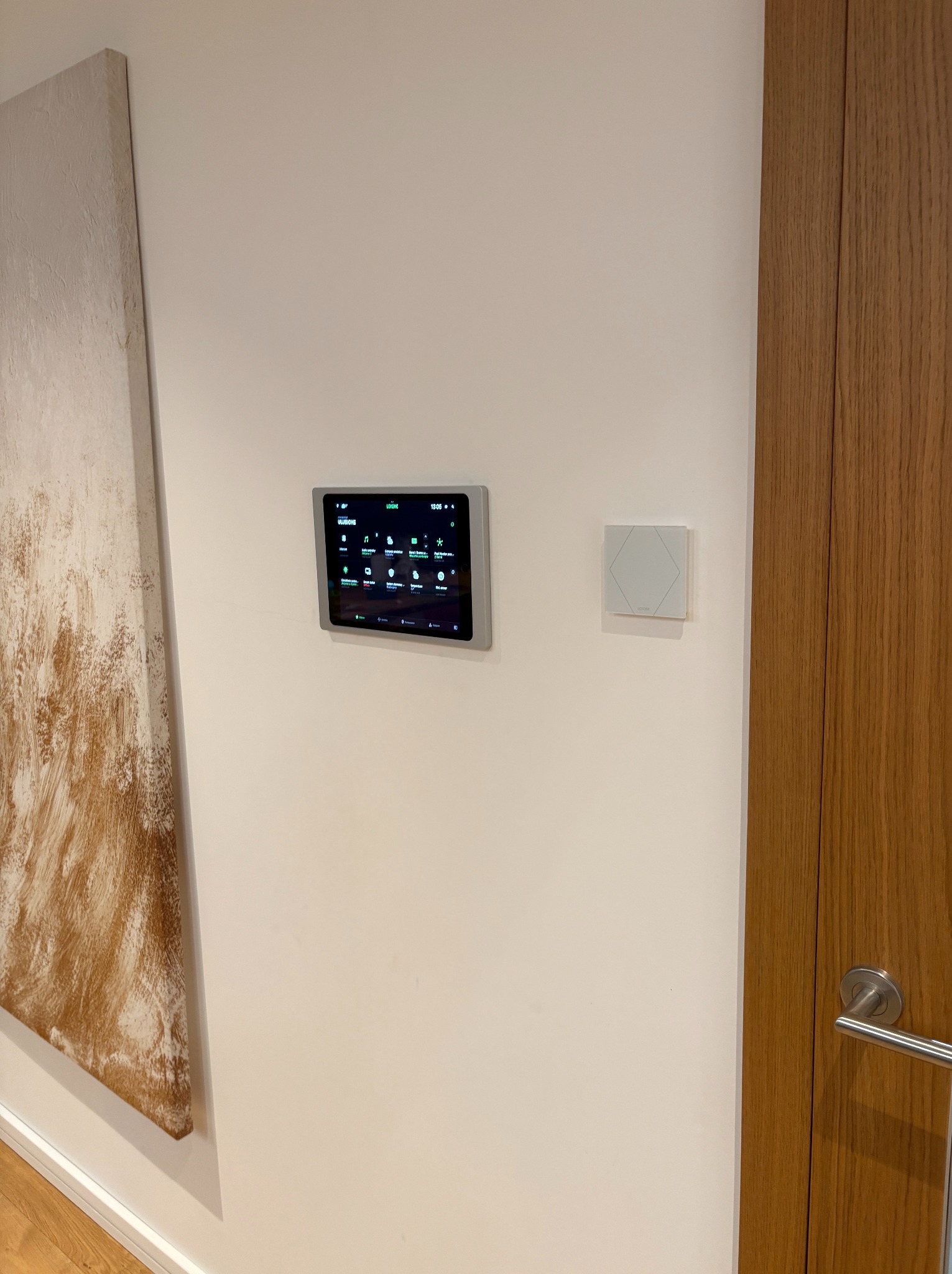 Panel sterowania inteligentnym domem zamontowany na ścianie, obok obrazu i drzwi z drewnianą okleiną.