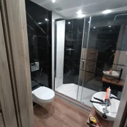 Nowoczesna łazienka z czarnymi płytkami imitującymi marmur, kabiną prysznicową typu walk-in z białymi płytkami wewnątrz, wiszącą toaletą i umywalką nablatową z narzędziami i silikonem na blacie...