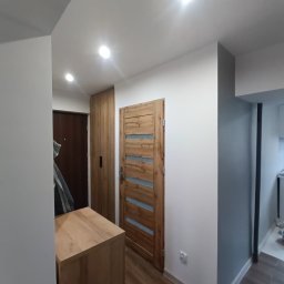 Kamińscy Ireneusz Kamiński - Wnętrze przedpokoju po remoncie: drewniane drzwi wewnętrzne z przeszkleniami, szafa w zabudowie, komoda, ściany pomalowane na biało i szaro, podłoga z paneli imitujących drewno.