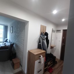 Kamińscy Ireneusz Kamiński - Przedpokój z szafką na buty z siedziskiem, wieszakiem na ubrania i lustrem, widok na salon z dekoracyjną ścianą z imitacji kamienia.