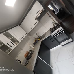 Kamińscy Ireneusz Kamiński - Nowoczesna kuchnia z białymi szafkami górnymi z przeszklonymi elementami i czarnymi uchwytami, szarym blatem, czarnymi szafkami dolnymi, białą marmurową podłogą i widocznym fragmentem salonu w tle.