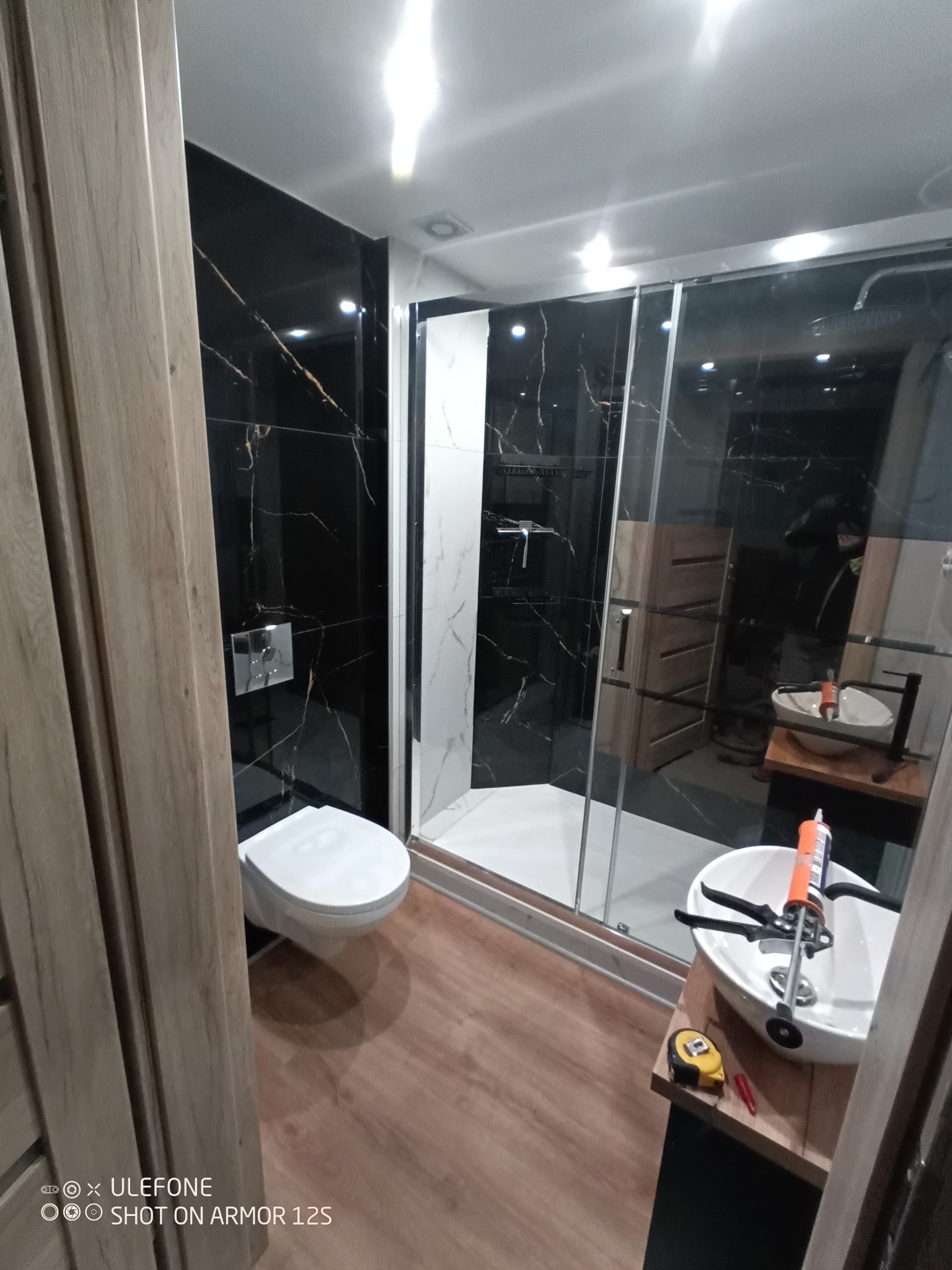 Nowoczesna łazienka z czarnymi płytkami imitującymi marmur, kabiną prysznicową typu walk-in z białymi płytkami wewnątrz, wiszącą toaletą i umywalką nablatową z narzędziami i silikonem na blacie...