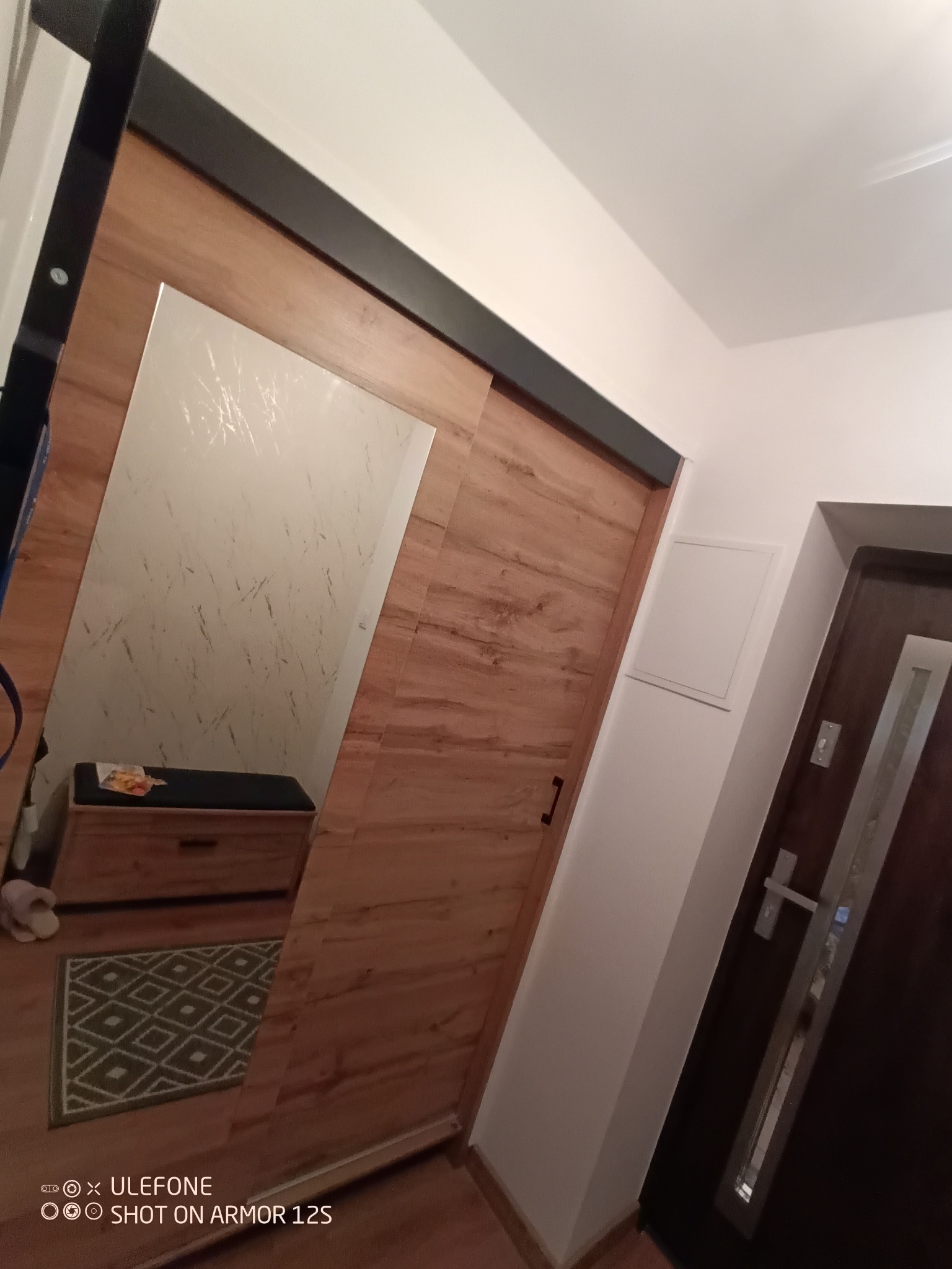 Drewniane drzwi przesuwne z czarną prowadnicą i lustrem na całej powierzchni, odbijające fragment przedpokoju z pufą i dywanem w geometryczny wzór.