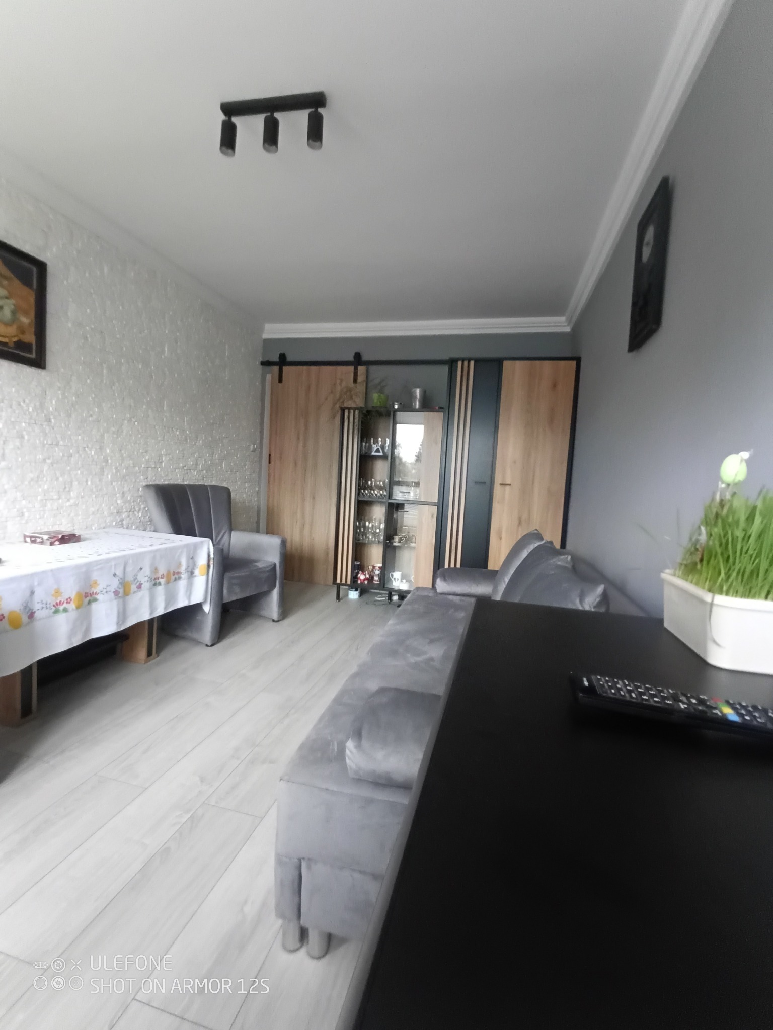 Nowoczesny salon z szarą sofą, fotelem i stołem z dekoracyjnym obrusem, ściana z białej cegły, drewniane drzwi przesuwne i regał z przeszkleniami.