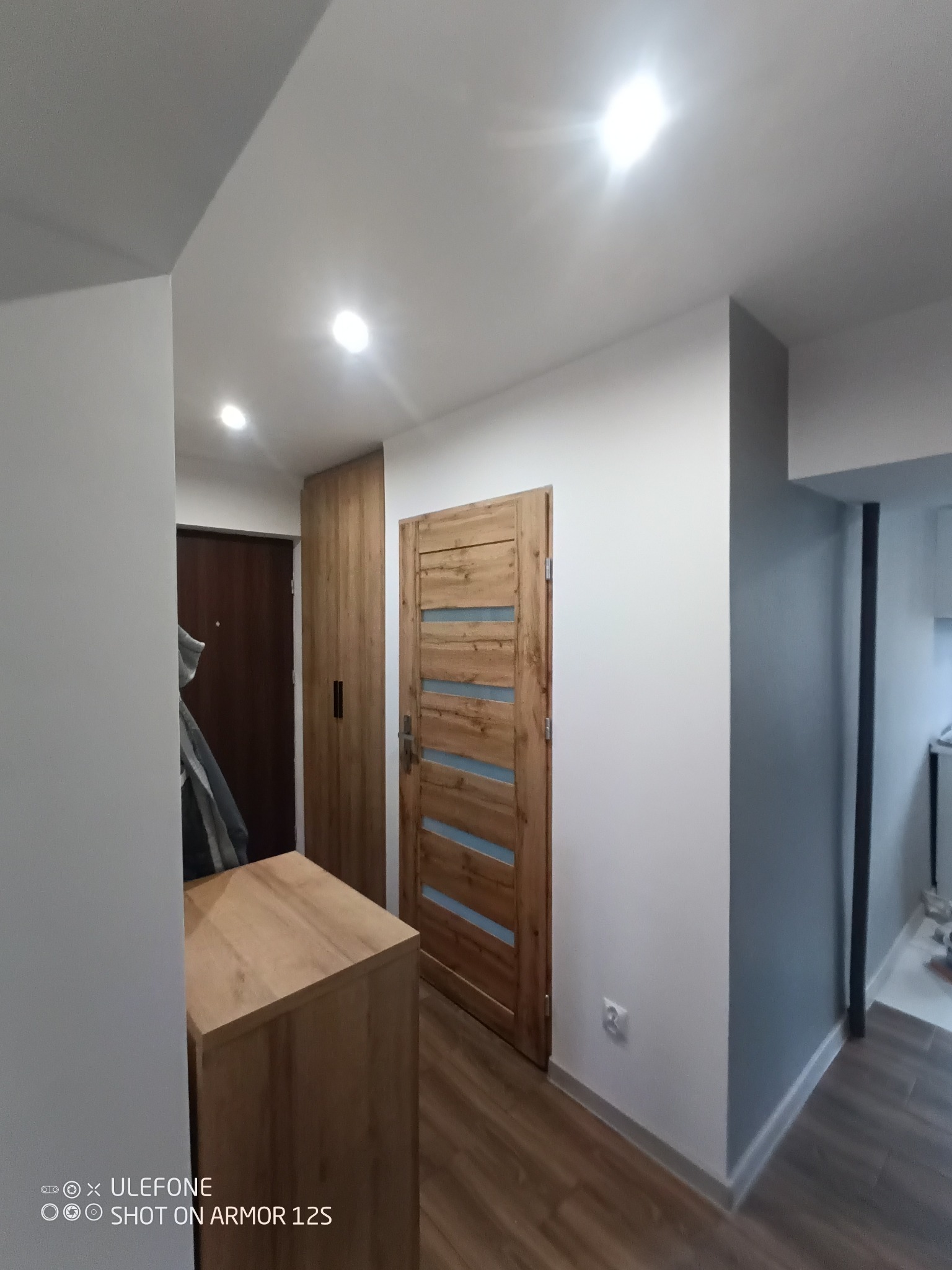 Wnętrze przedpokoju po remoncie: drewniane drzwi wewnętrzne z przeszkleniami, szafa w zabudowie, komoda, ściany pomalowane na biało i szaro, podłoga z paneli imitujących drewno.