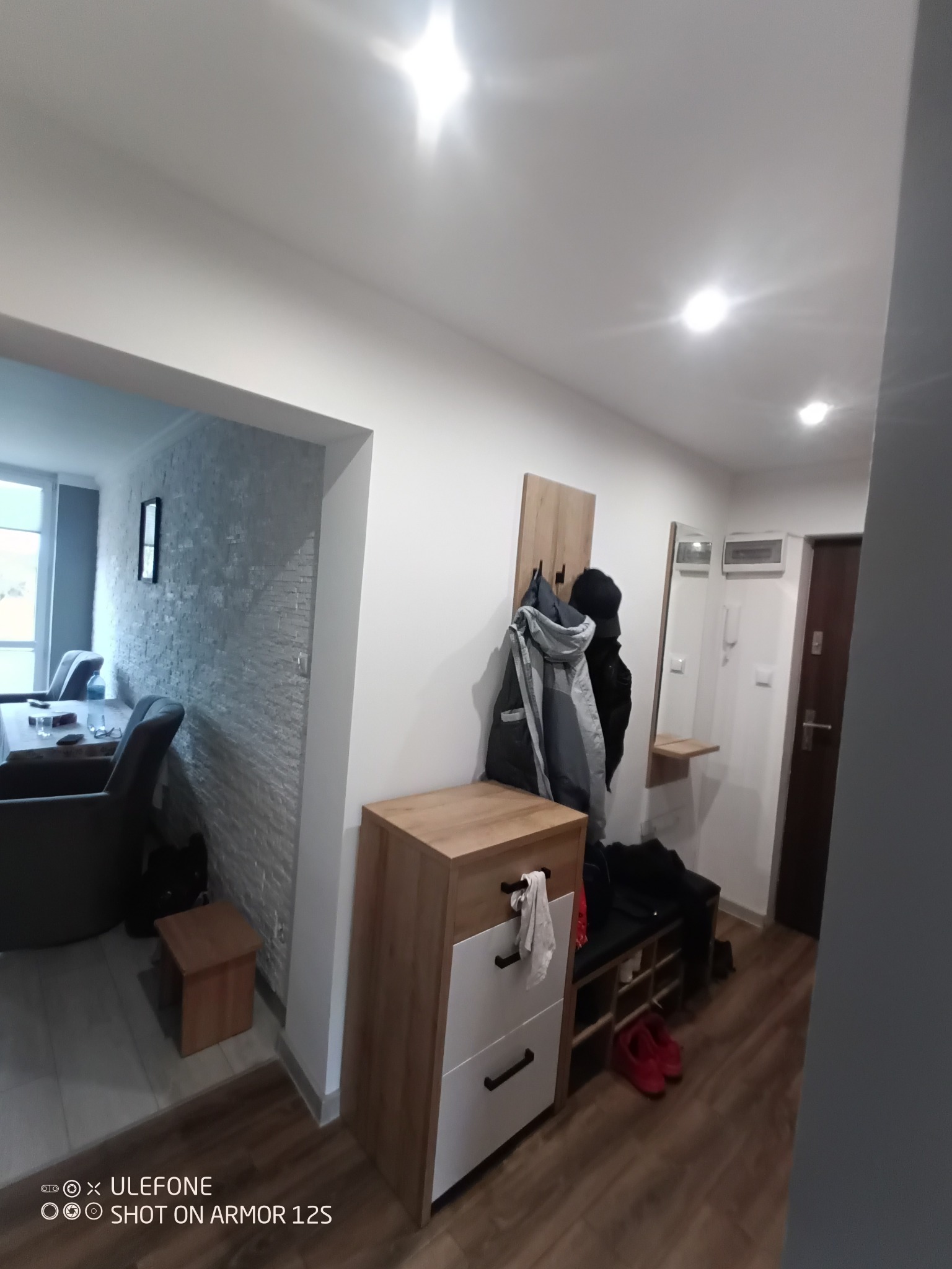 Przedpokój z szafką na buty z siedziskiem, wieszakiem na ubrania i lustrem, widok na salon z dekoracyjną ścianą z imitacji kamienia.