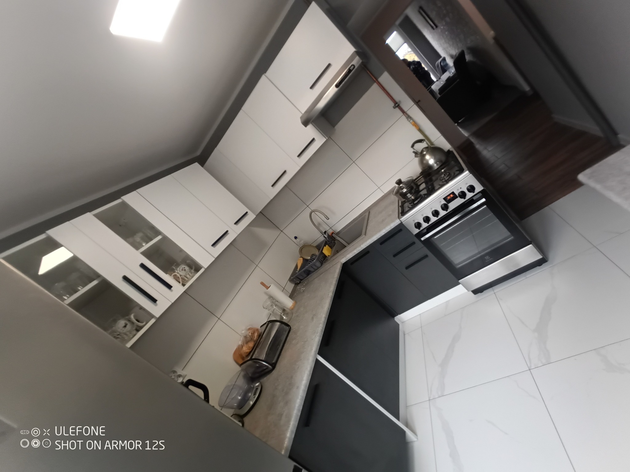 Nowoczesna kuchnia z białymi szafkami górnymi z przeszklonymi elementami i czarnymi uchwytami, szarym blatem, czarnymi szafkami dolnymi, białą marmurową podłogą i widocznym fragmentem salonu w tle.