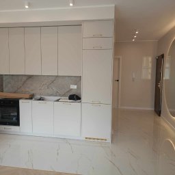 VIK-BUD VIKTOR SYDORCHUK - Wykończona kuchnia z białymi szafkami z delikatnymi złotymi uchwytami, marmurowym blatem i płytkami podłogowymi imitującymi marmur, widok na przedpokój z okrągłym lustrem z podświetleniem LED.