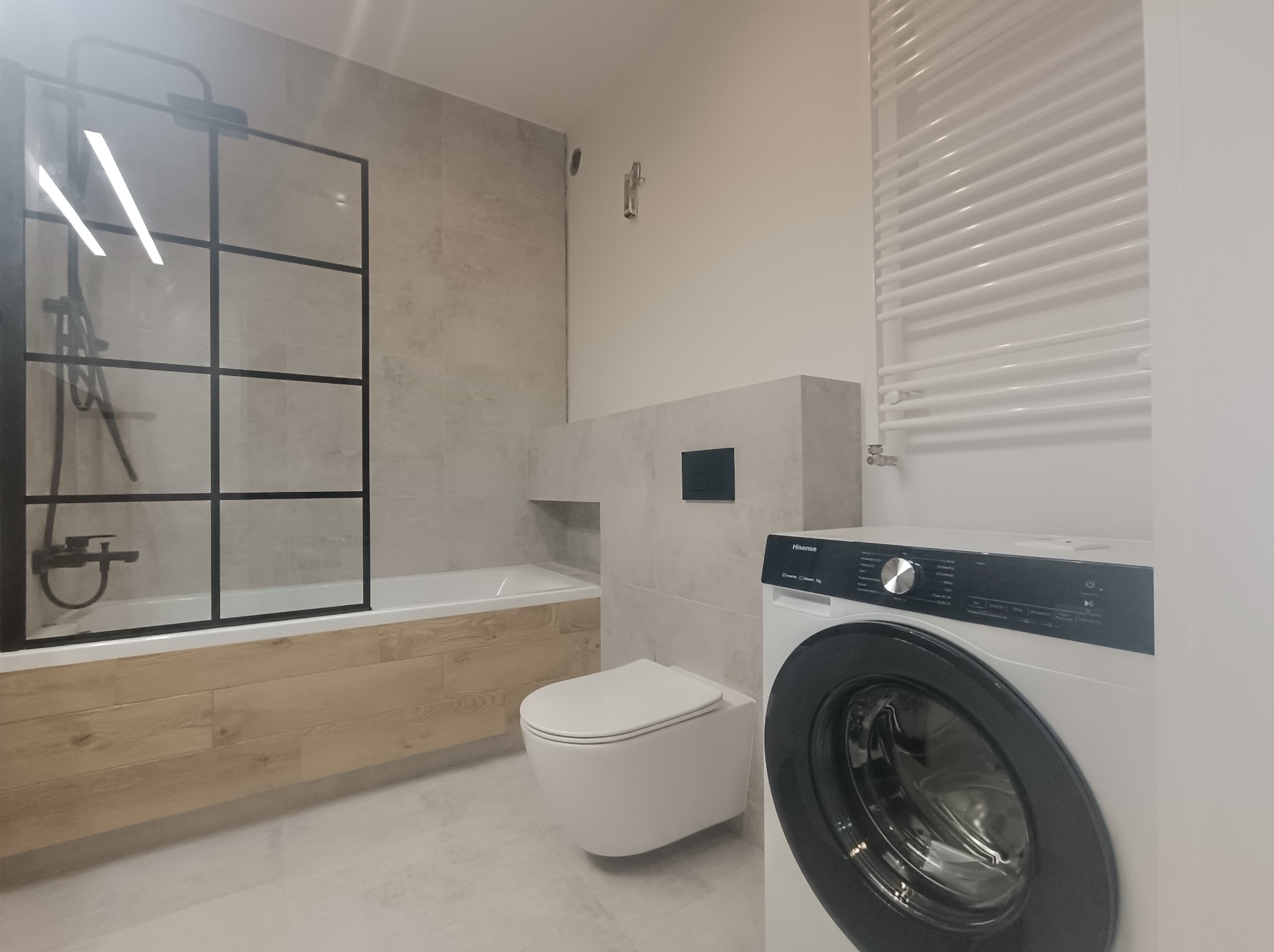 Nowoczesna łazienka z kabiną prysznicową typu walk-in z czarnymi profilami, wiszącą toaletą, grzejnikiem drabinkowym i pralką w jasnej kolorystyce.