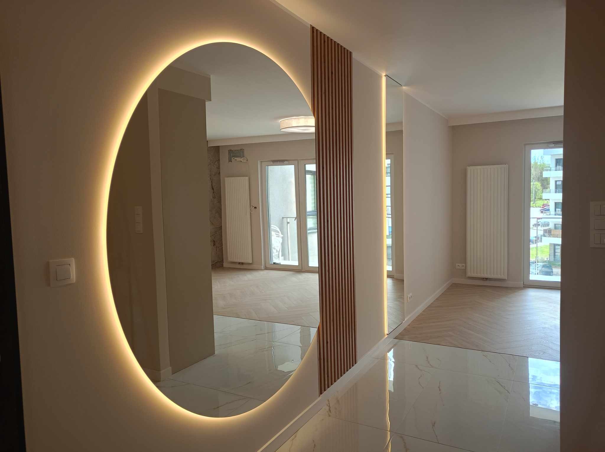 Nowoczesne wnętrze z okrągłym lustrem podświetlanym LED, drewnianymi lamelami na ścianie, błyszczącą podłogą z białego marmuru i parkietem w jodełkę w tle.