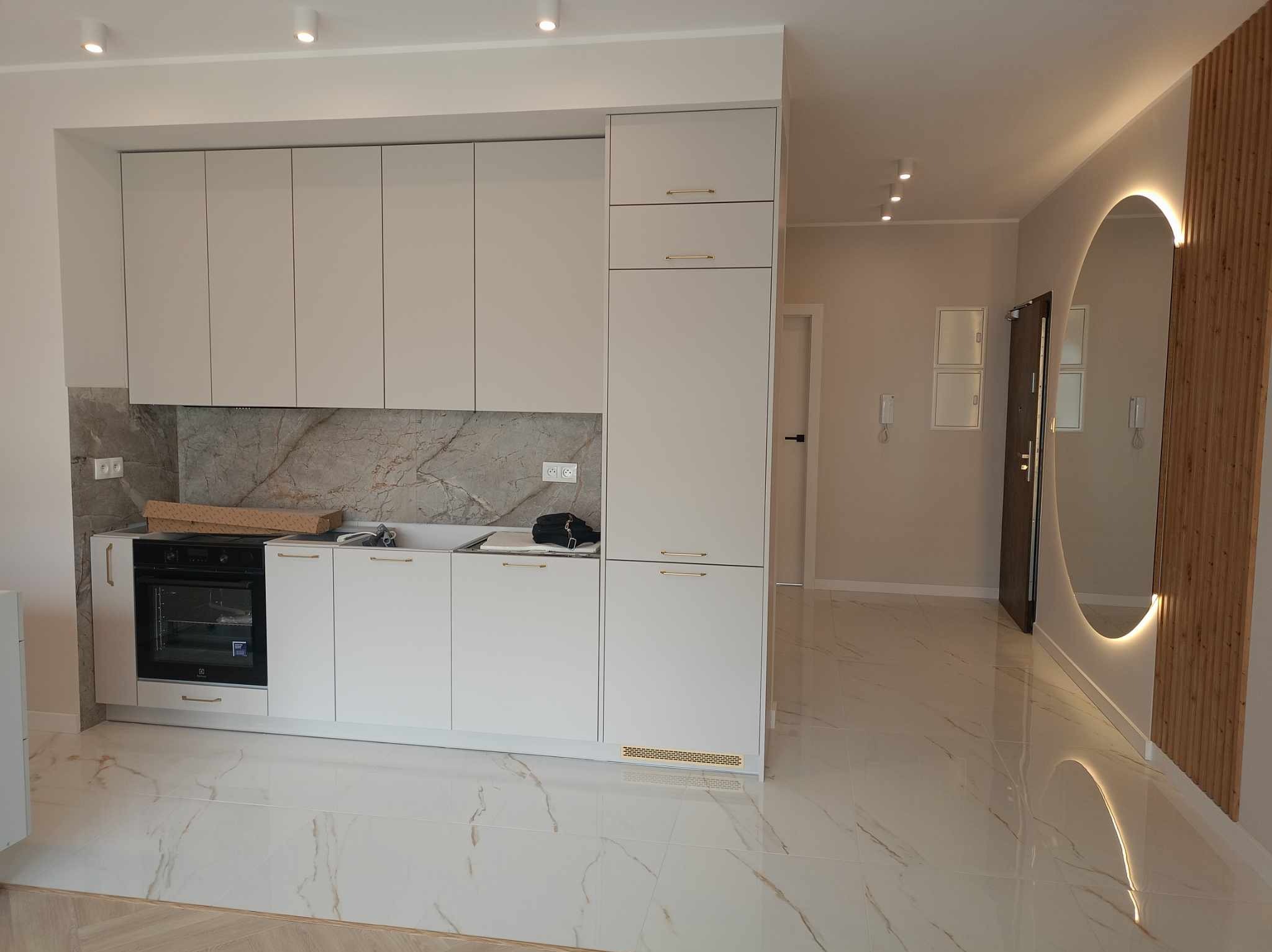 Wykończona kuchnia z białymi szafkami z delikatnymi złotymi uchwytami, marmurowym blatem i płytkami podłogowymi imitującymi marmur, widok na przedpokój z okrągłym lustrem z podświetleniem LED.