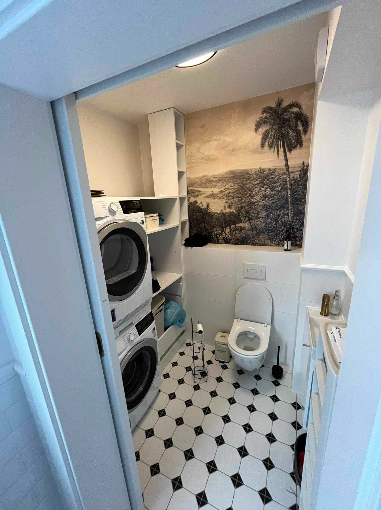 Wnętrze łazienki z toaletą, umywalką, pralką i suszarką w zabudowie, z czarno-białą podłogą w karo i tapetą z motywem tropikalnym na ścianie.