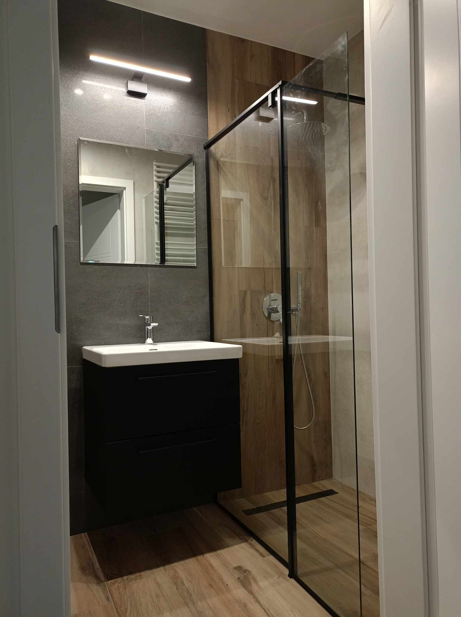 Nowoczesna łazienka z kabiną prysznicową typu walk-in z czarnymi profilami, umywalką nablatową na czarnej szafce, szarą glazurą i drewnopodobnymi płytkami na ścianach w strefie prysznica.