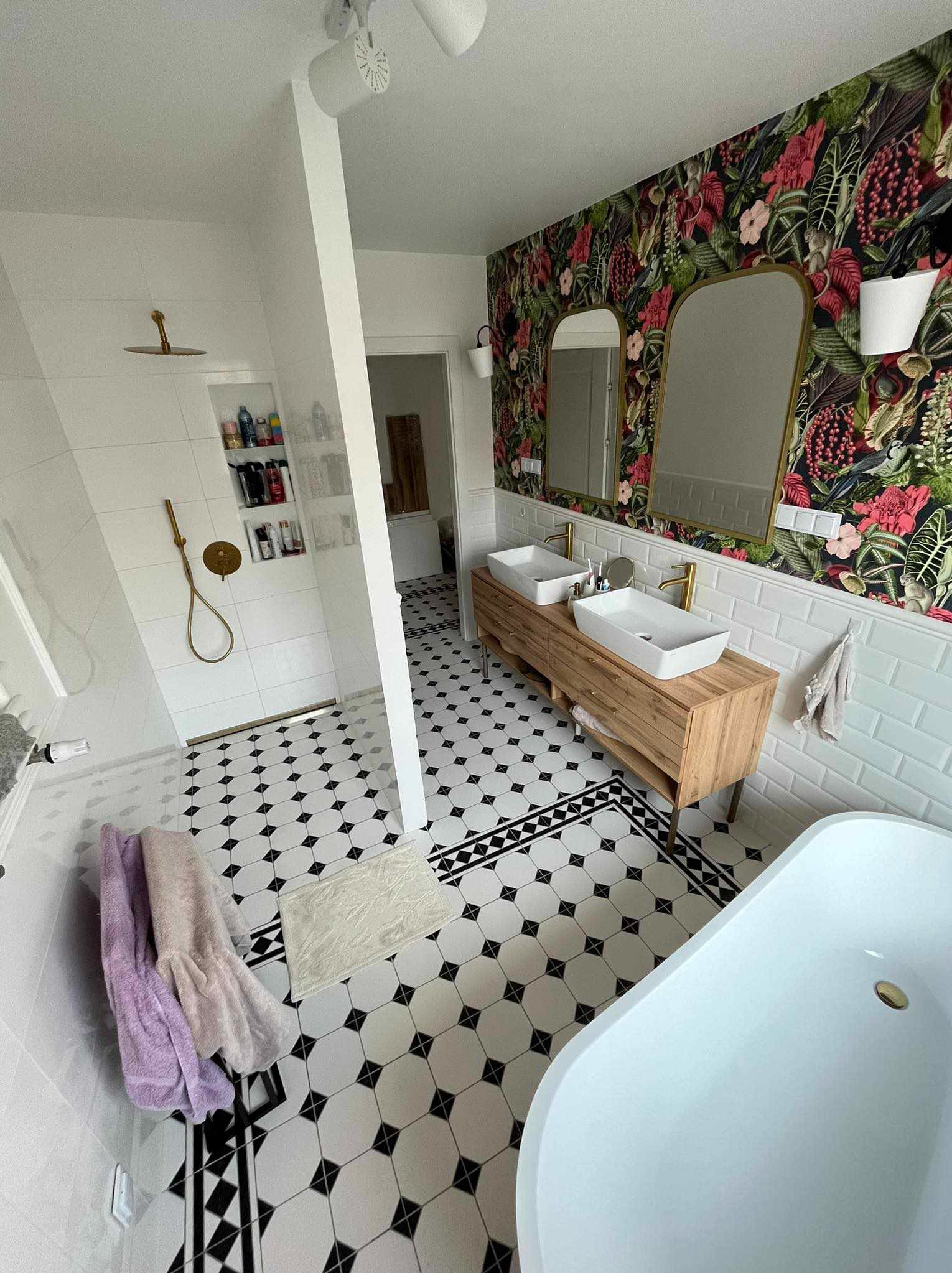 Nowoczesna łazienka z wanną wolnostojącą, dwiema umywalkami nablatowymi na drewnianej szafce, czarno-białą podłogą w karo, kabiną prysznicową typu walk-in z złotą armaturą i ścianą z tapetą...