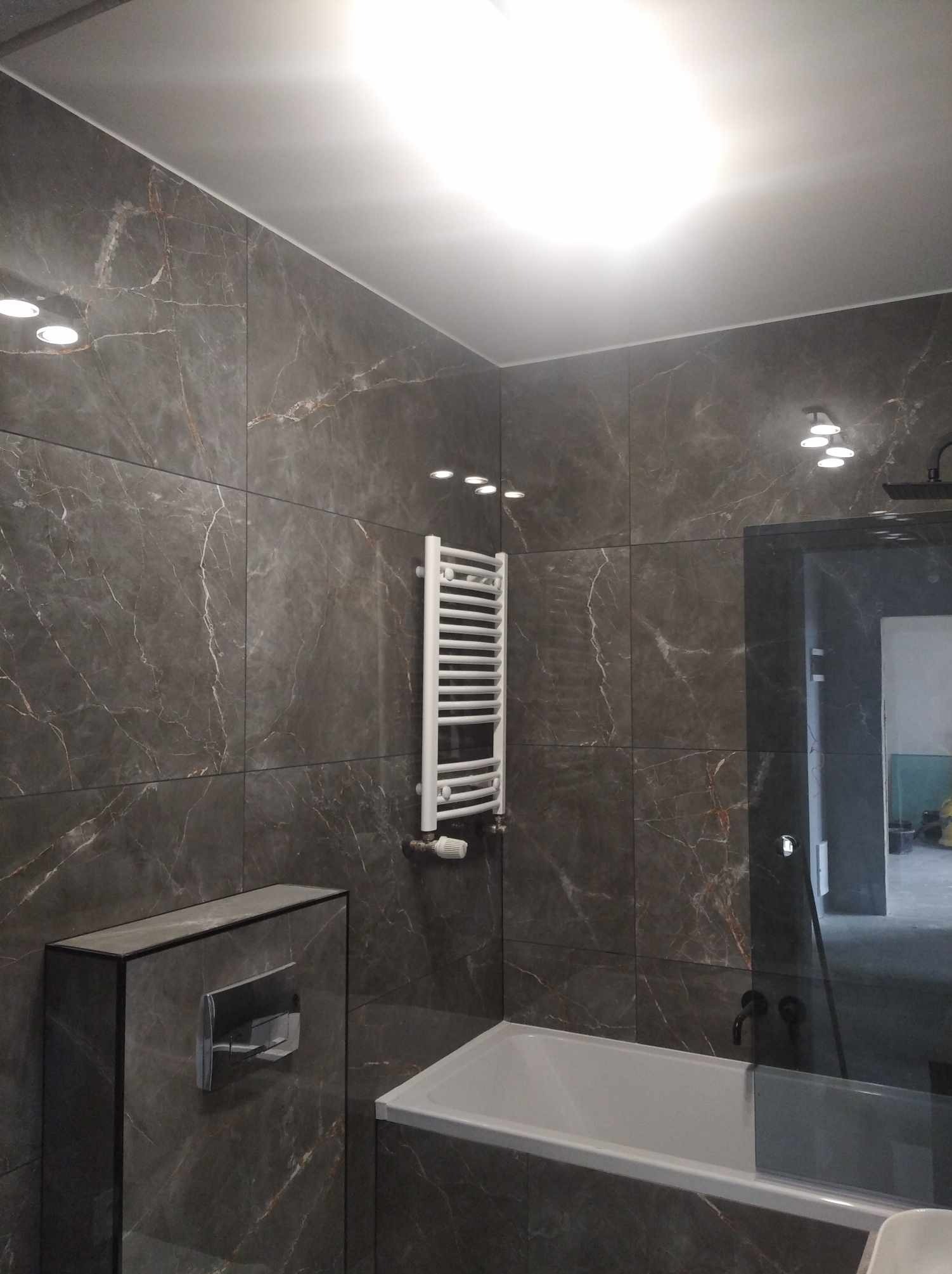 Nowoczesna łazienka wykończona płytkami imitującymi marmur, z białą wanną, grzejnikiem drabinkowym i podtynkową spłuczką WC.