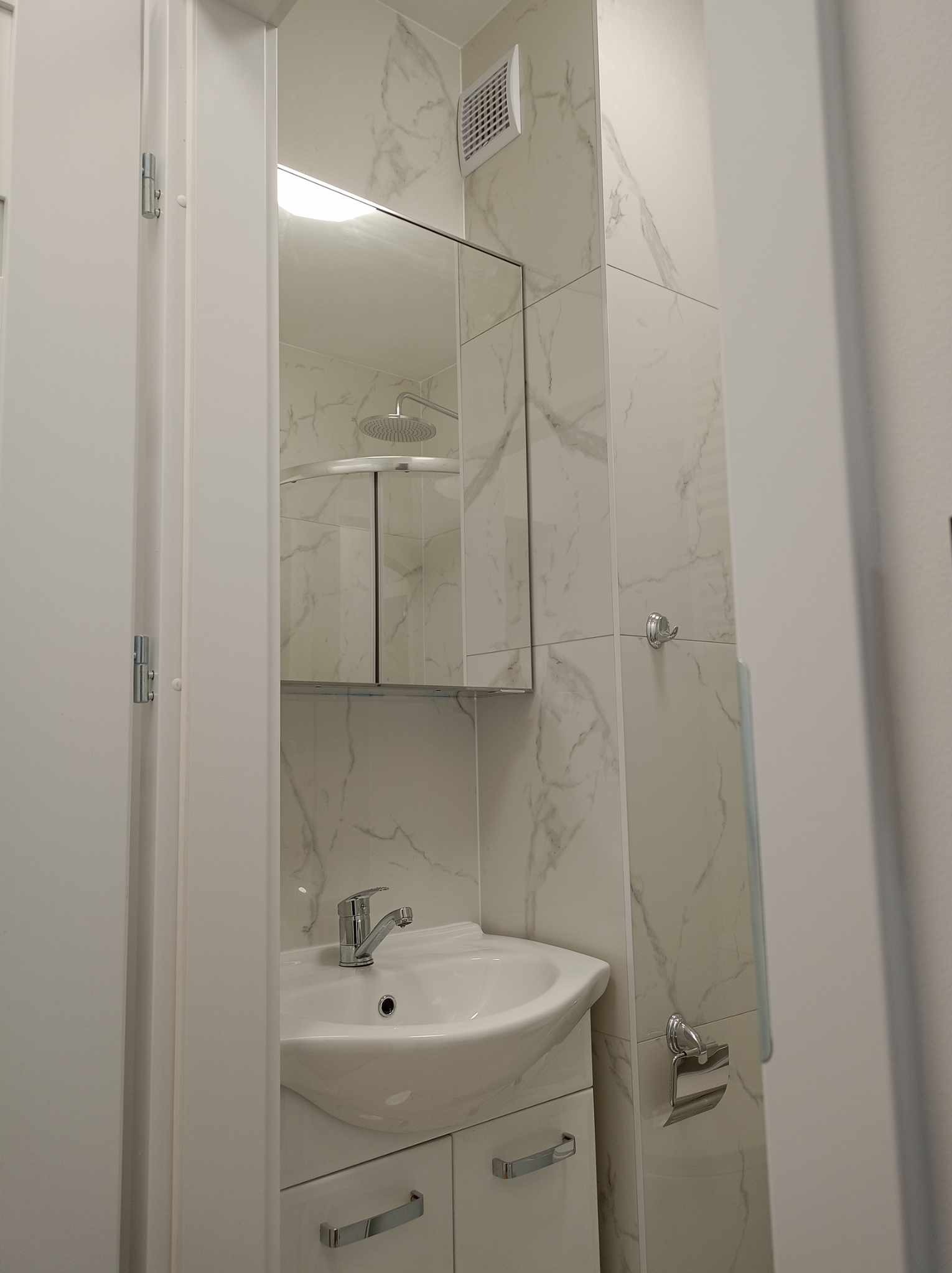 Umywalka z szafką i lustrem w wykończonej łazience, ściany wyłożone białymi płytkami imitującymi marmur, widoczny fragment kabiny prysznicowej.